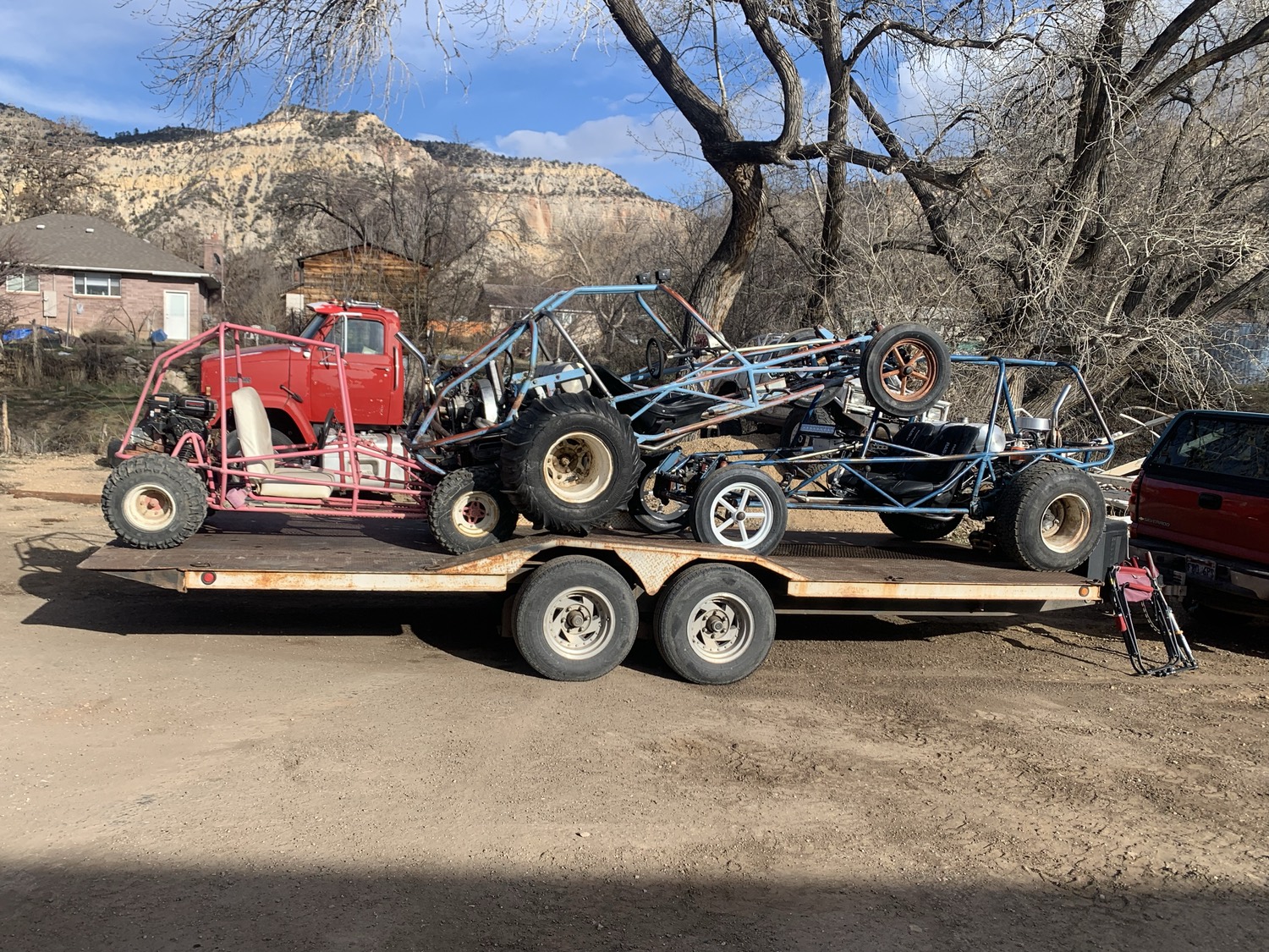 Three sandrail buggies loaded on a flatbed trailer below the cliffs, an old red cabover truck behind