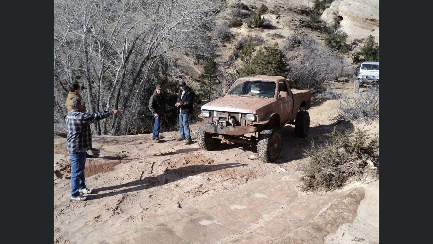 A mud-caked lifted mini truck crawling a slickrock ledge while the crew spots the line